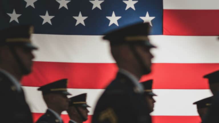 Blurred silhouettes of soldiers in uniform walking in front of a large, in-focus American flag.