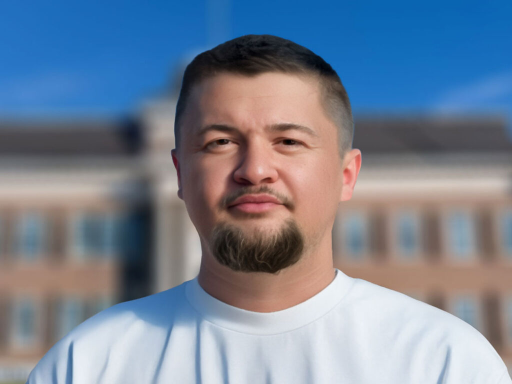 Close-up portrait of a man with short dark hair and facial hair, wearing a white T-shirt, outdoors with a blurred building behind him and blue sky.