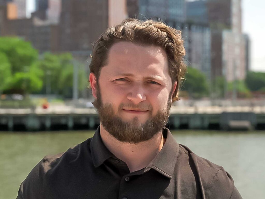 Portrait of a man with a beard and brown hair, wearing a dark shirt, standing outdoors by a river with city buildings in the background.