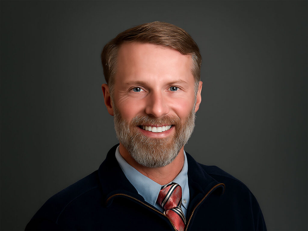 Smiling man with a gray beard wearing a blue shirt and striped tie against a dark background.
